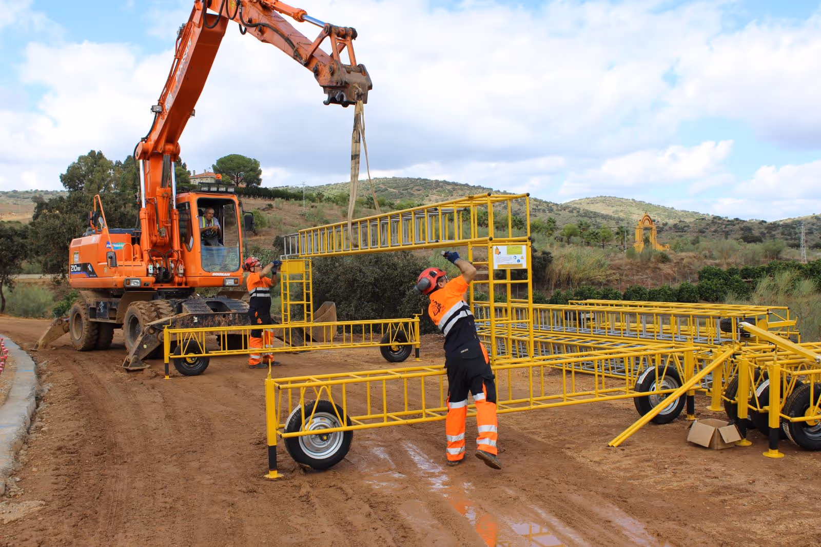Trabajadores de construcción con ropa de seguridad naranja ensamblan una estructura metálica amarilla con la ayuda de una excavadora en un terreno de tierra.
