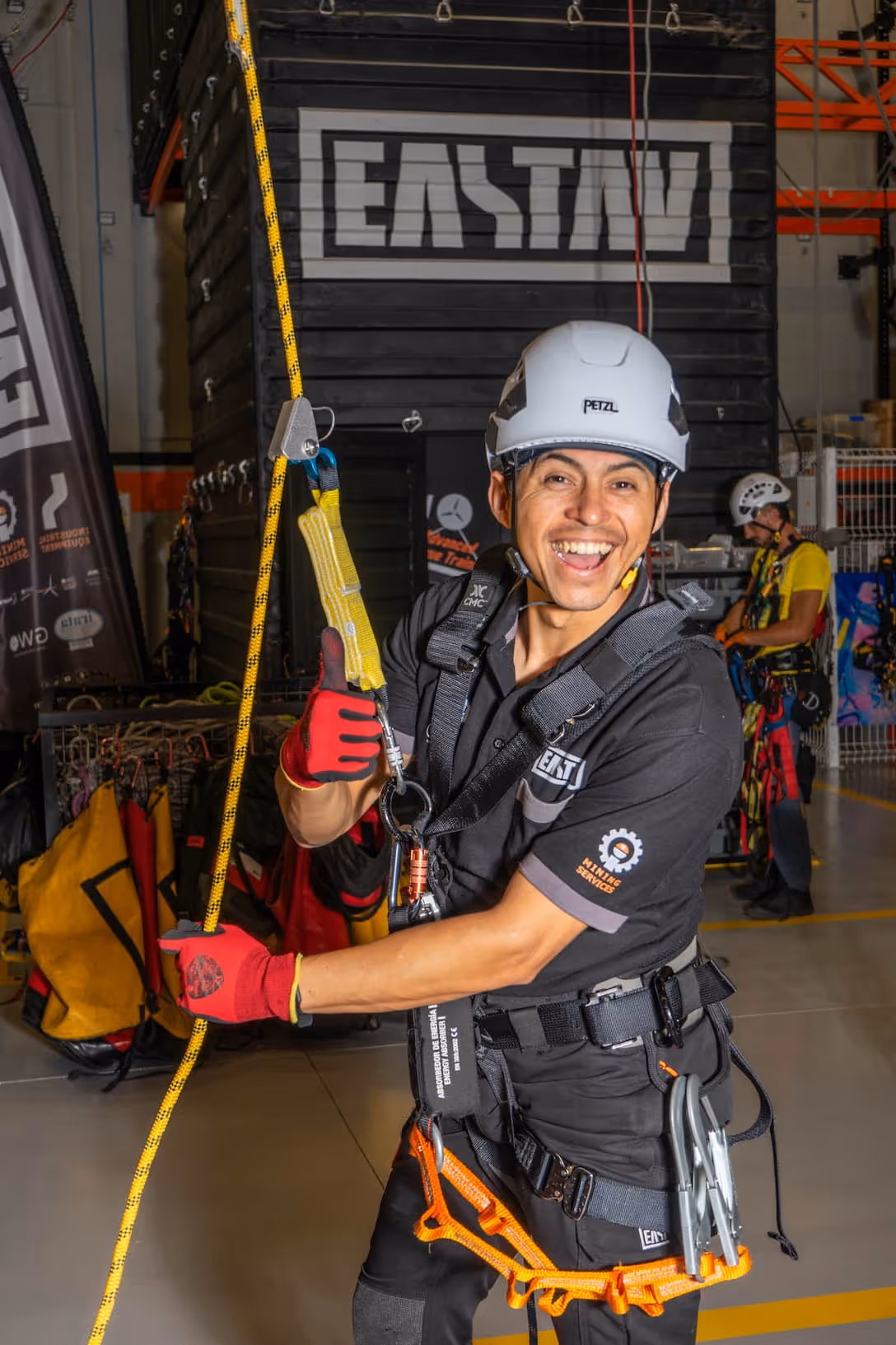 Trabajador sonriente con arnés y casco blanco sostiene una cuerda amarilla en un taller industrial.