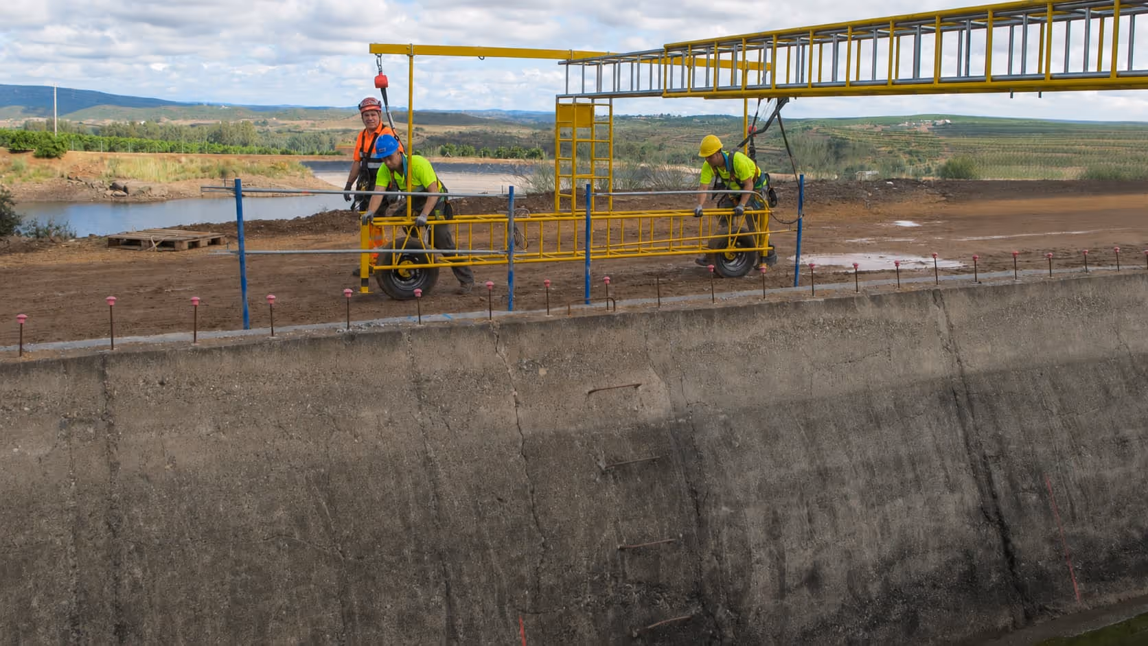 Tres trabajadores con arneses en una plataforma amarilla suspendida sobre un muro de concreto alto.