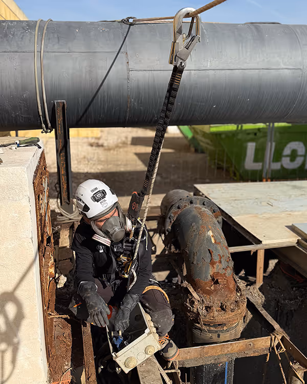 Trabajador con casco y mascarilla realizando mantenimiento en tubería oxidada en zona industrial.