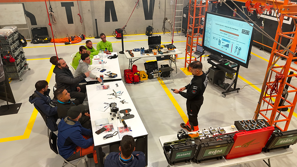 Grupo de personas en taller observan drones y equipos de tecnología en mesas amarillas.