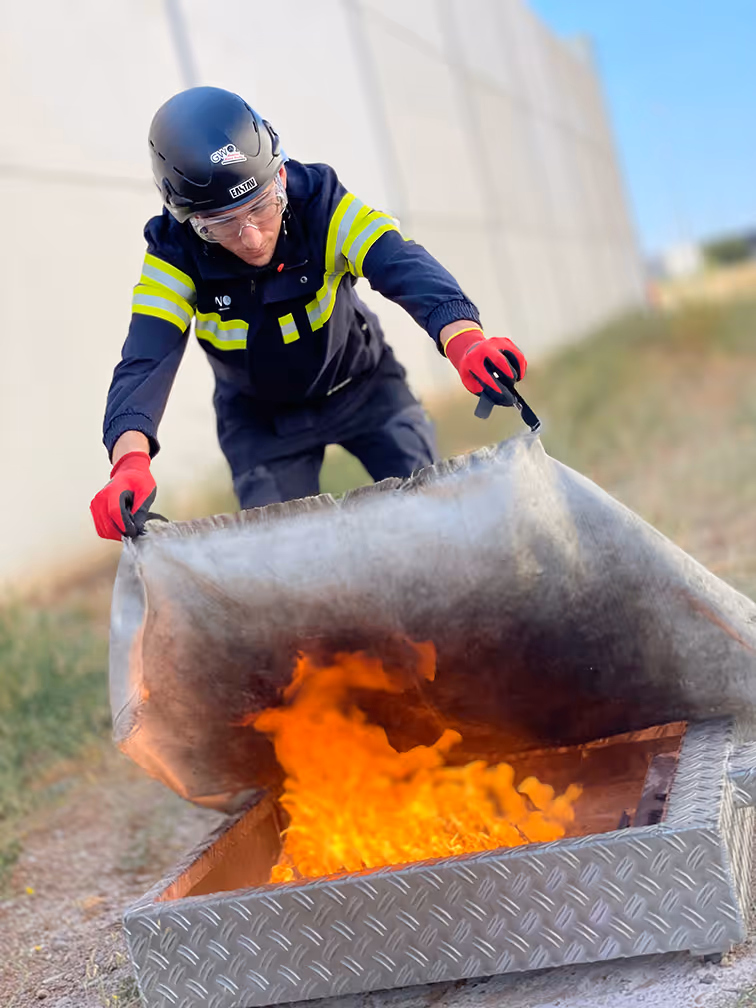 Bombero con equipo de protección sofoca fuego usando manta metálica especializada.