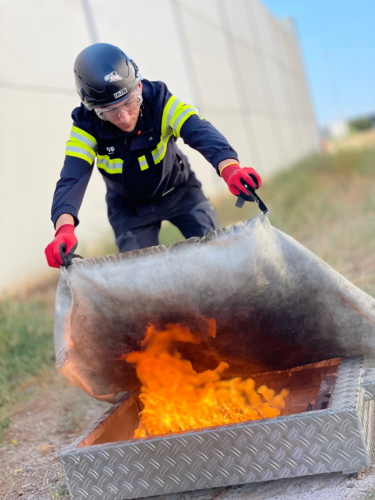 Bombero con equipo de protección sofoca fuego usando manta metálica especializada.