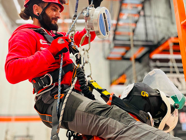 Trabajador en rojo con arnés de seguridad realizando tareas en altura con equipo profesional.