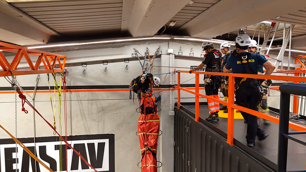 Instructores con cascos en plataforma naranja supervisan entrenamiento de rescate en altura con equipos profesionales.