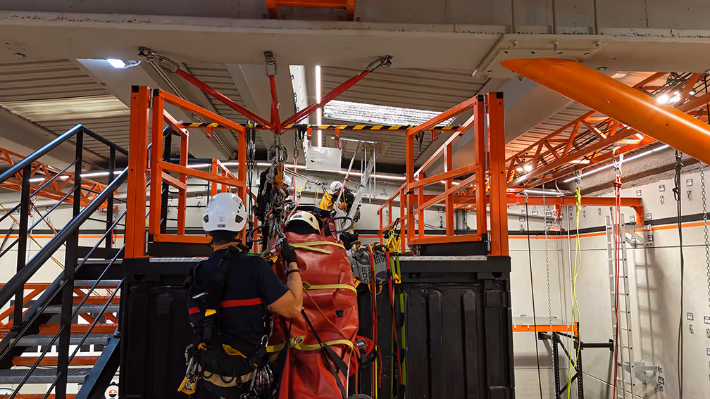Trabajadores en arnés de seguridad practican rescate en altura con estructura metálica roja.