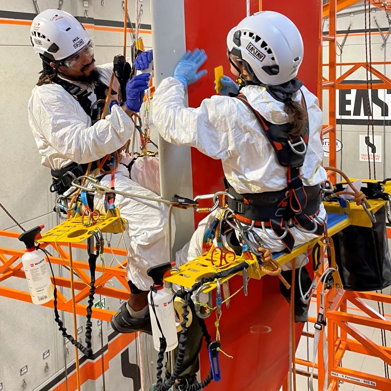 Dos trabajadores de altura con cascos blancos, arneses de seguridad y guantes azules trabajan en una estructura industrial roja.