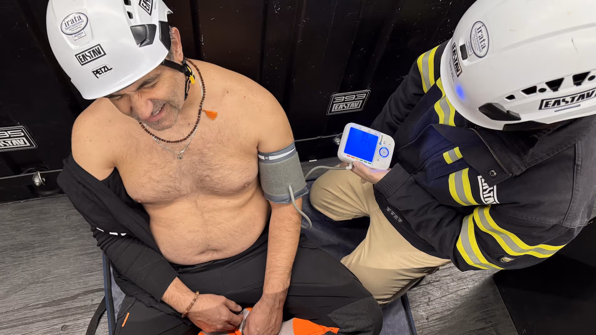 Two climbers wearing safety helmets examine a blue climbing route-finding device together indoors.
