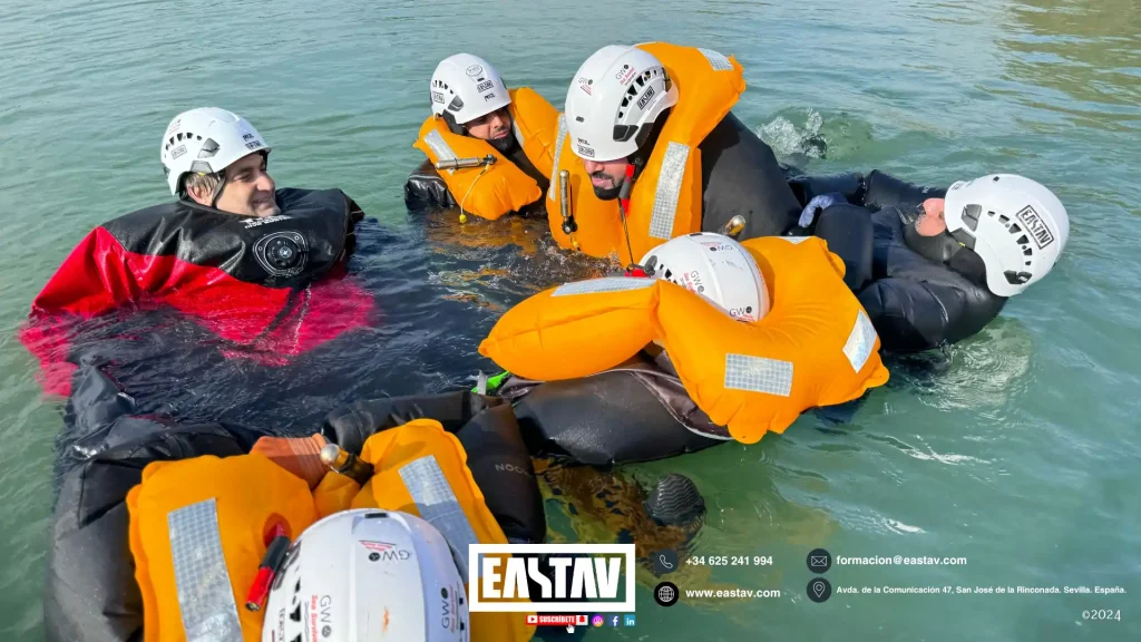 Grupo de personas en el agua usando cascos y chalecos salvavidas naranjas durante entrenamiento acuático.