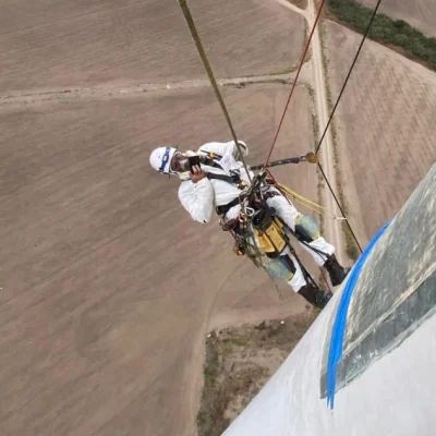 Worker suspended by ropes and harness descending from tall structure with safety equipment.