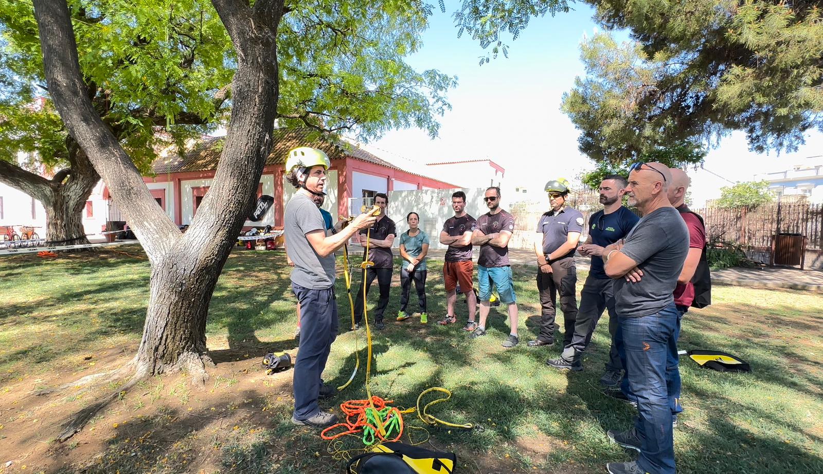 Arborist demonstrates tree climbing and safety equipment to a group of trainees outdoors.