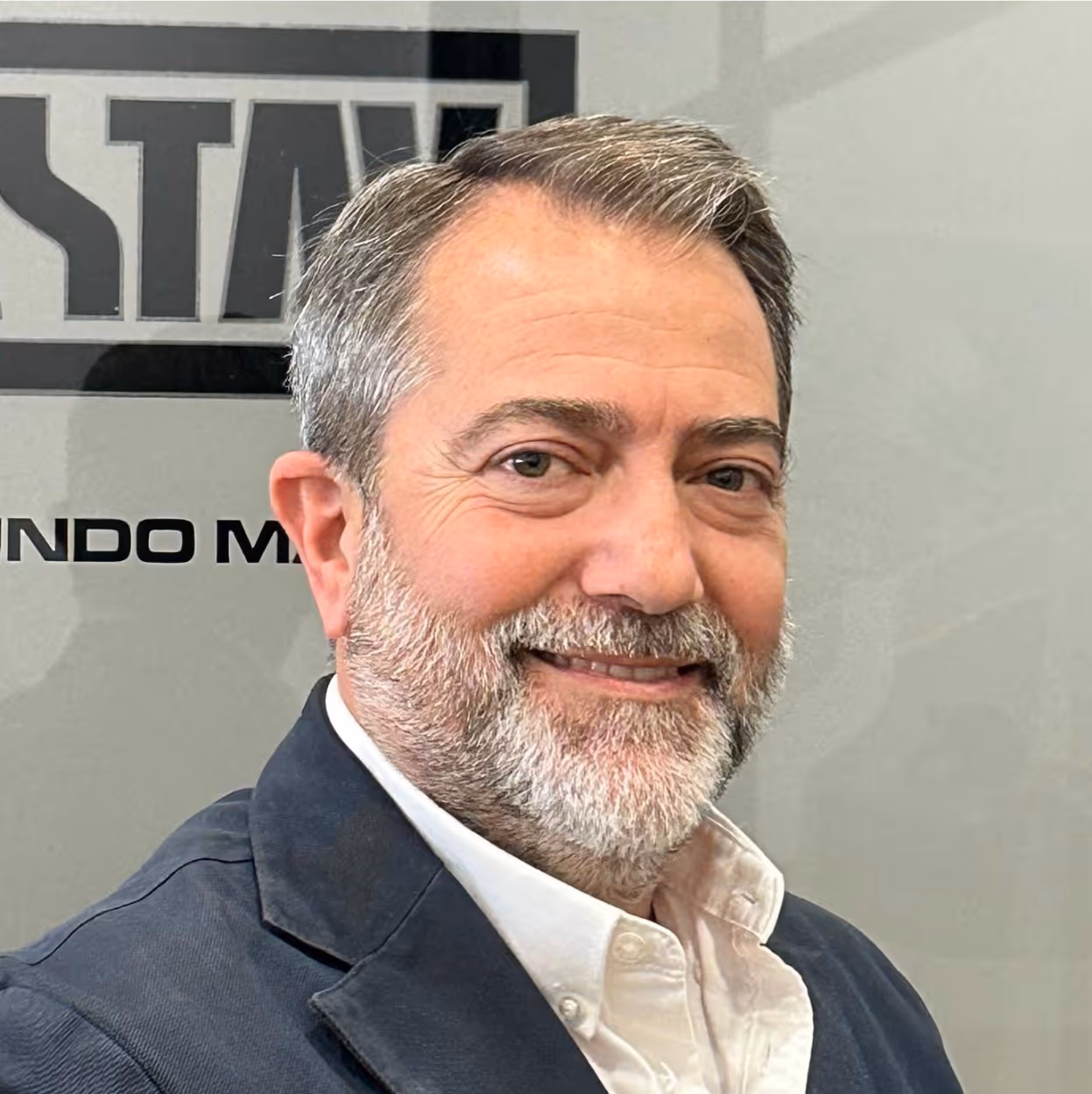 Hombre adulto con barba gris, chaqueta oscura y camisa blanca sonriendo a cámara.