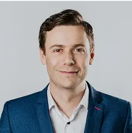 Portrait of a man in a blue suit jacket and white shirt, smiling at the camera against a light background.