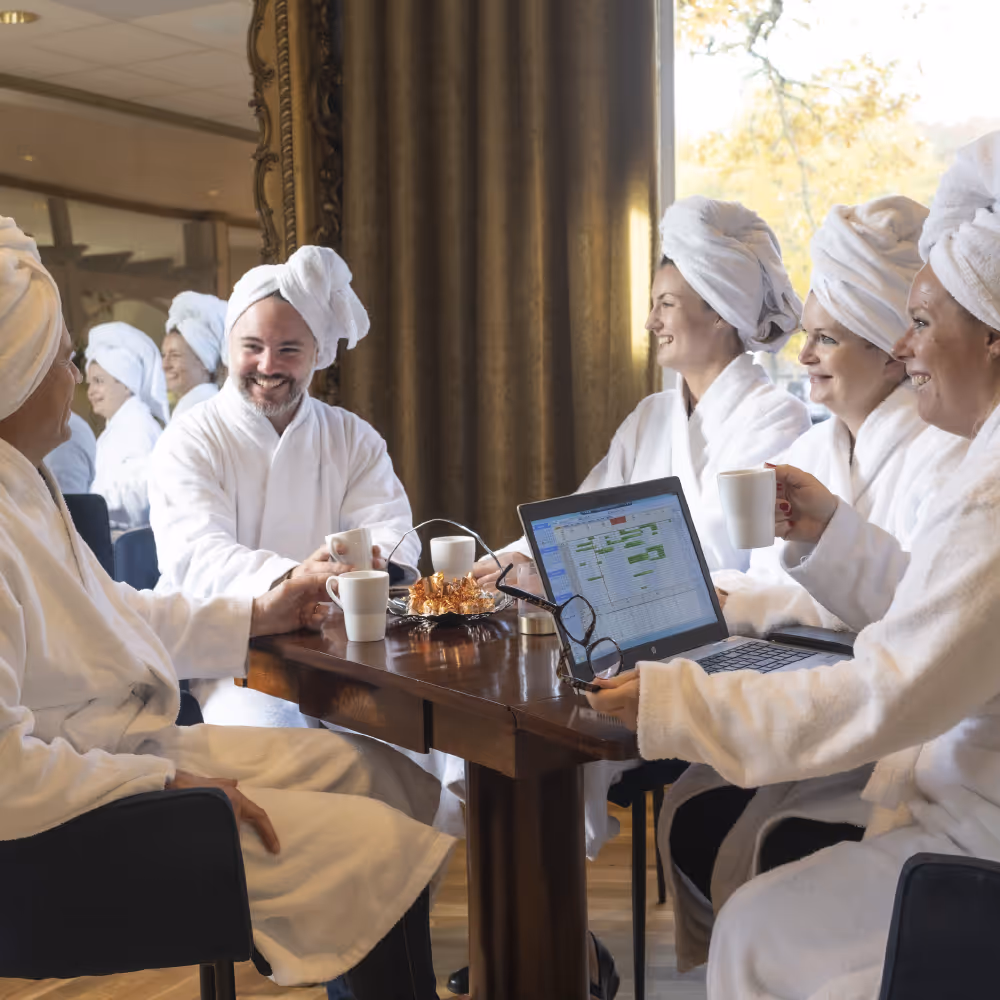 Personer i badrockar har ett avslappnat möte med kaffe och laptop vid ett bord.