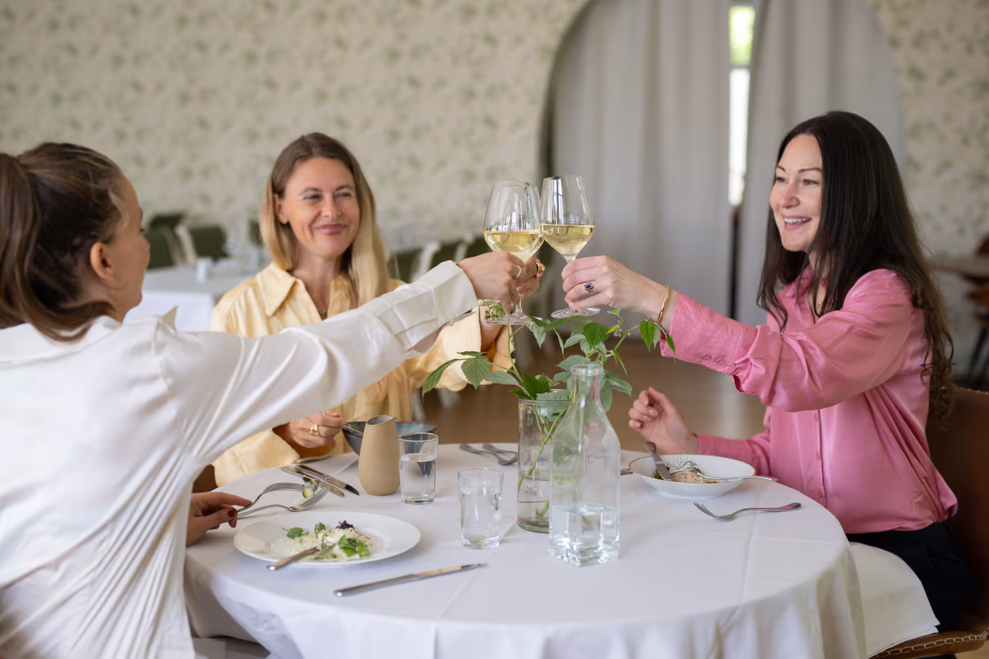 Tre kvinnor som skålar med vitvinsglas vid ett restaurangbord.