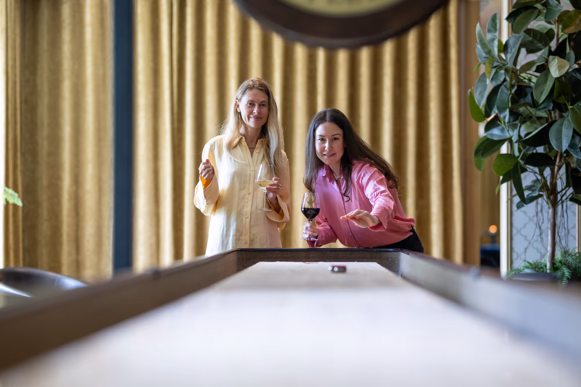 Två kvinnor spelar shuffleboard medan de njuter av vin i en elegant miljö.