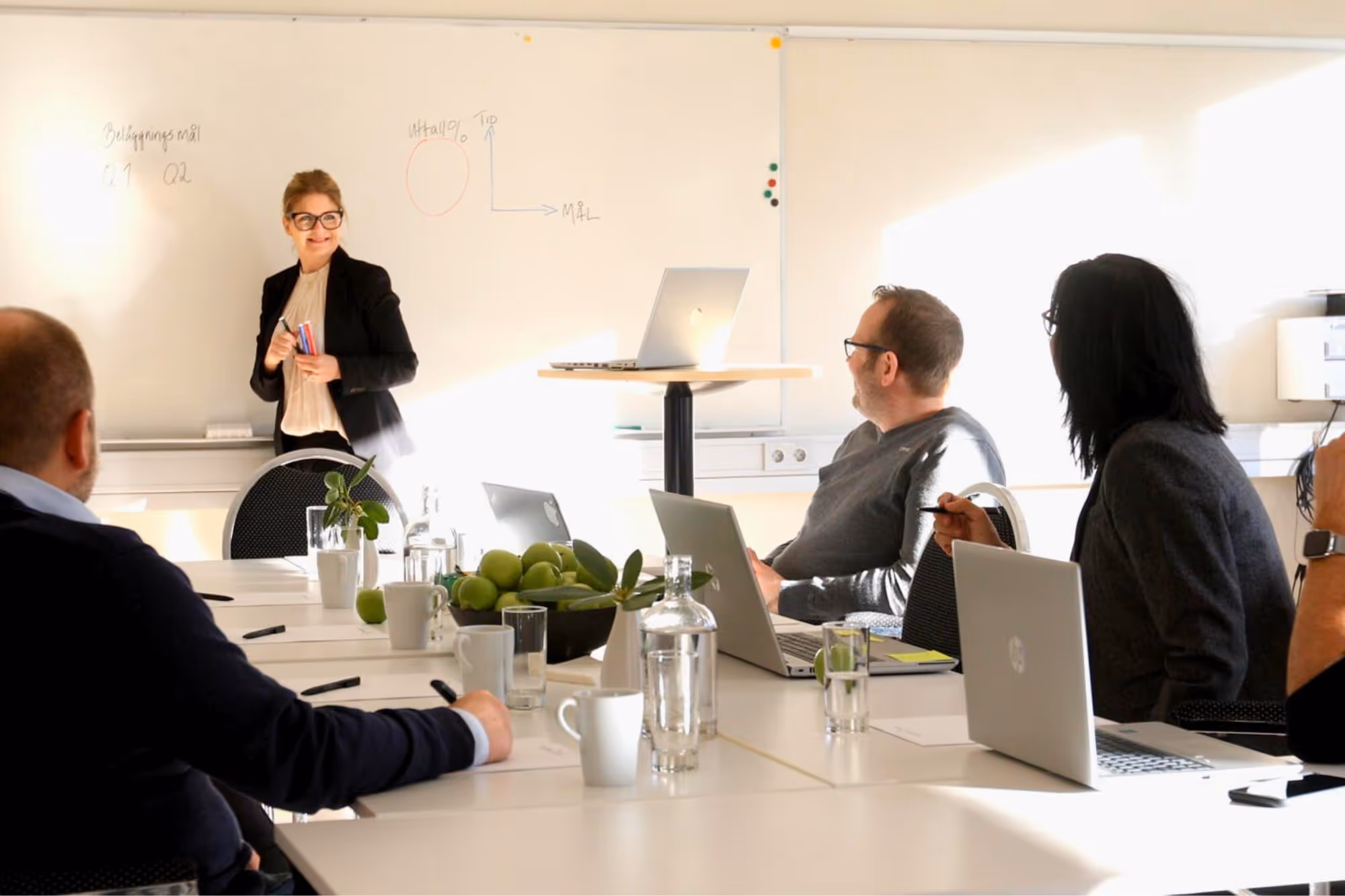 Person håller en presentation i ett modernt konferensrum, deltagare med laptops antecknar.