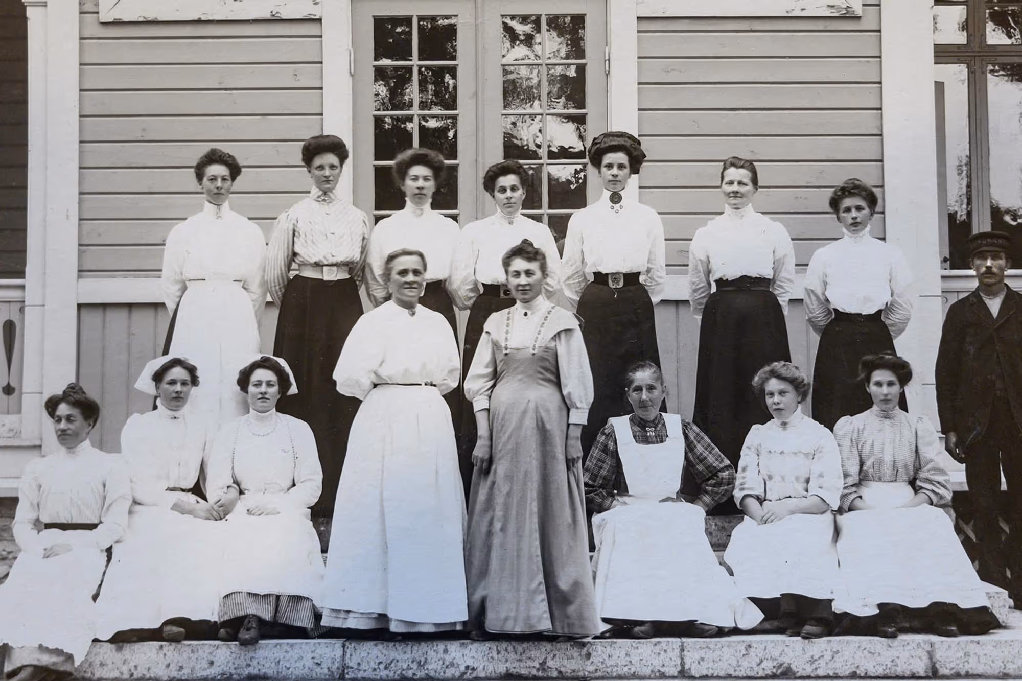 Gamla fotografi av grupp människor i tidstypiska kläder framför trähus.