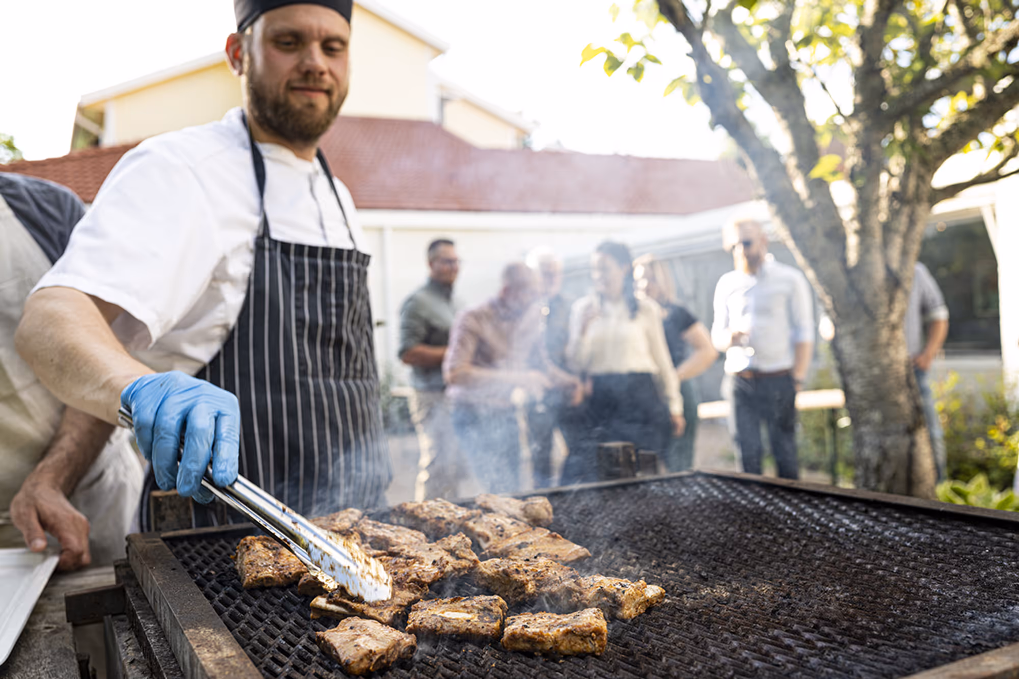 Man with randig förkläde och blå handskar grillar kött på en stor utomhusgrill med en grupp människor i bakgrunden.