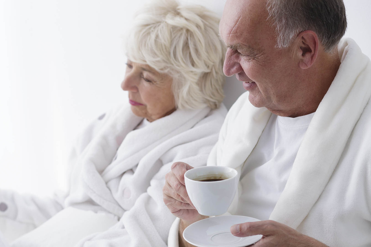 Äldre par i vita morgonrockar, mannen håller en kopp kaffe och ler.