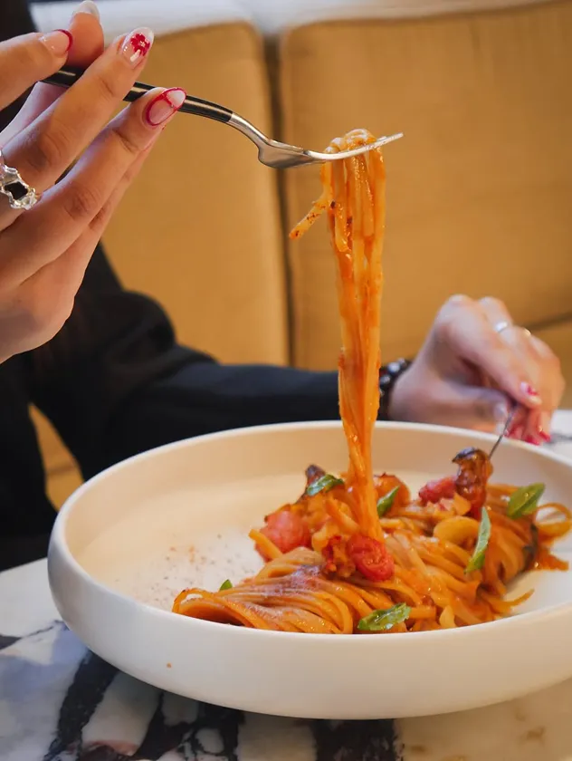 A person twirling long pasta with a fork from a white bowl, showing red nail polish.
