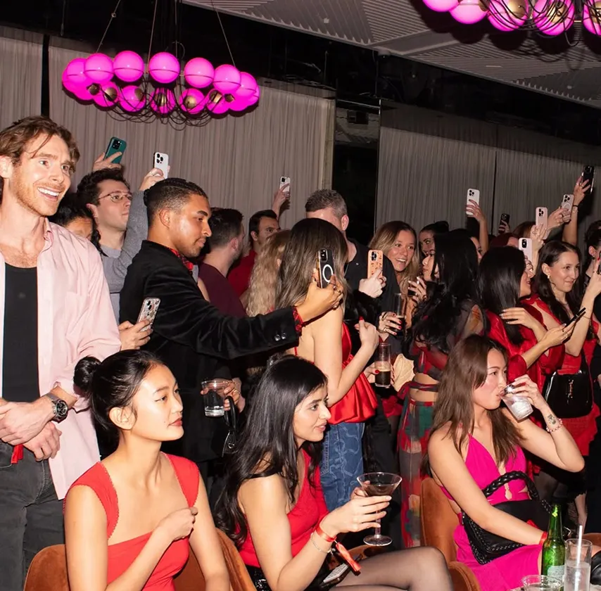 People socializing and taking photos at a lively event under pink lights.