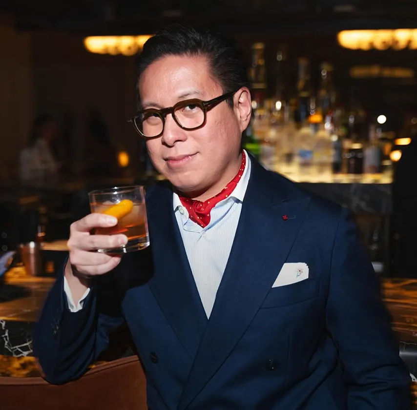 A man in a blue suit and red scarf holds a cocktail in a dimly lit bar.