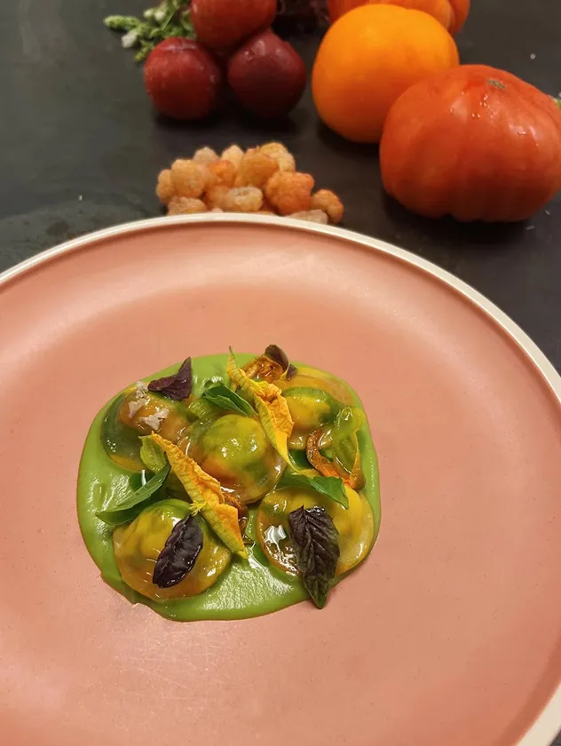 Gourmet pasta dish with green sauce on a pink plate, with fruits and vegetables in the background.
