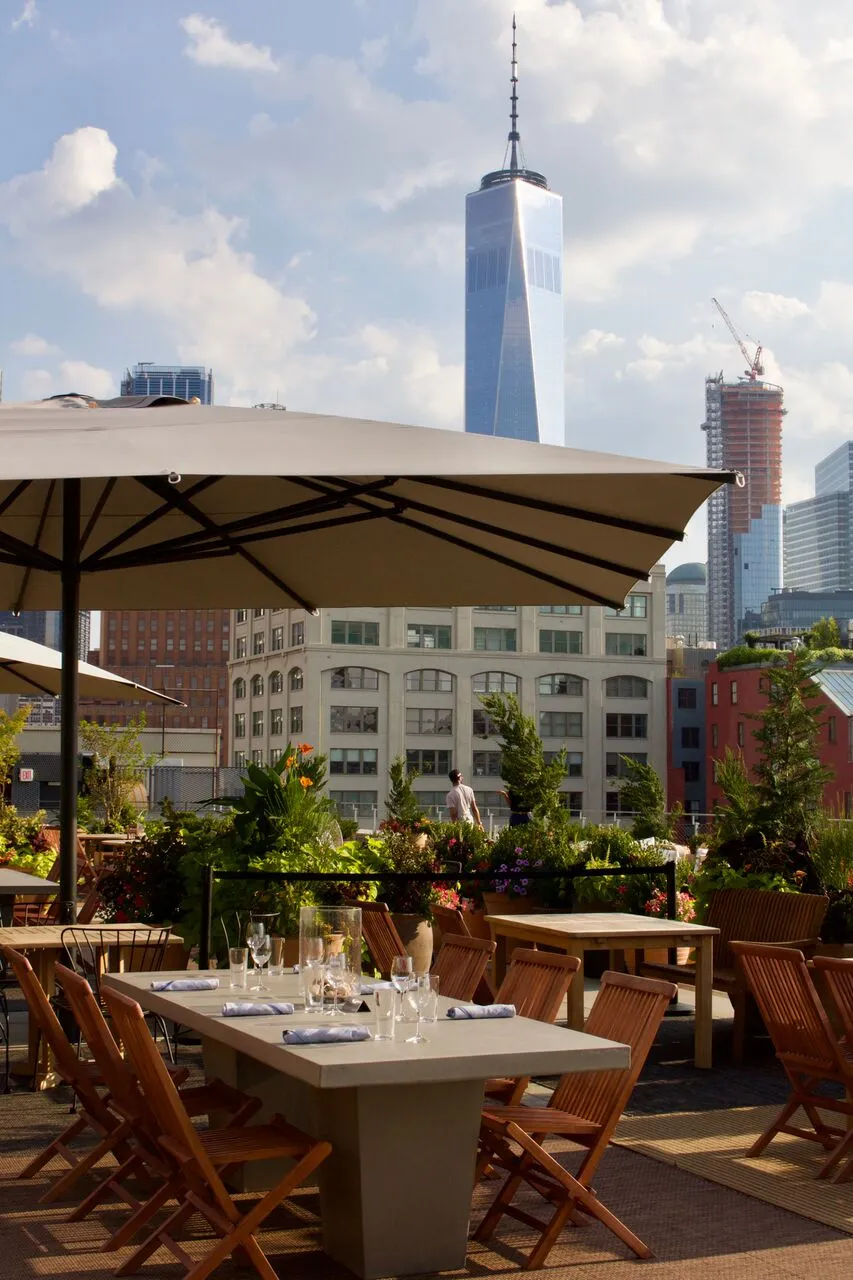An outdoor rooftop dining area with tables set under umbrellas, surrounded by lush greenery, offering a clear view of the iconic One World Trade Center skyscraper in downtown Manhattan.