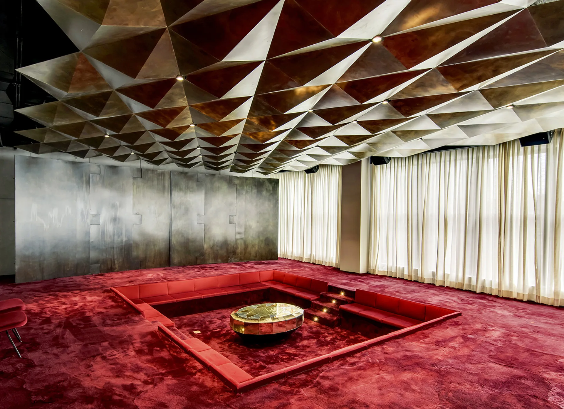A luxurious room with a striking red sunken seating area, gold triangular ceiling, and long white curtains.
