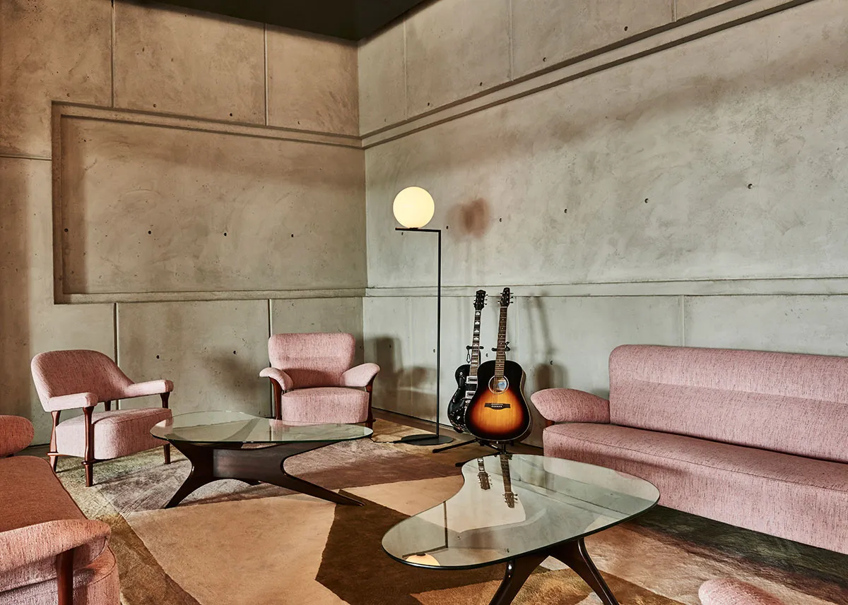 A stylish lounge area with pink sofas and armchairs, glass coffee tables, and two guitars by a globe lamp, set against concrete walls.