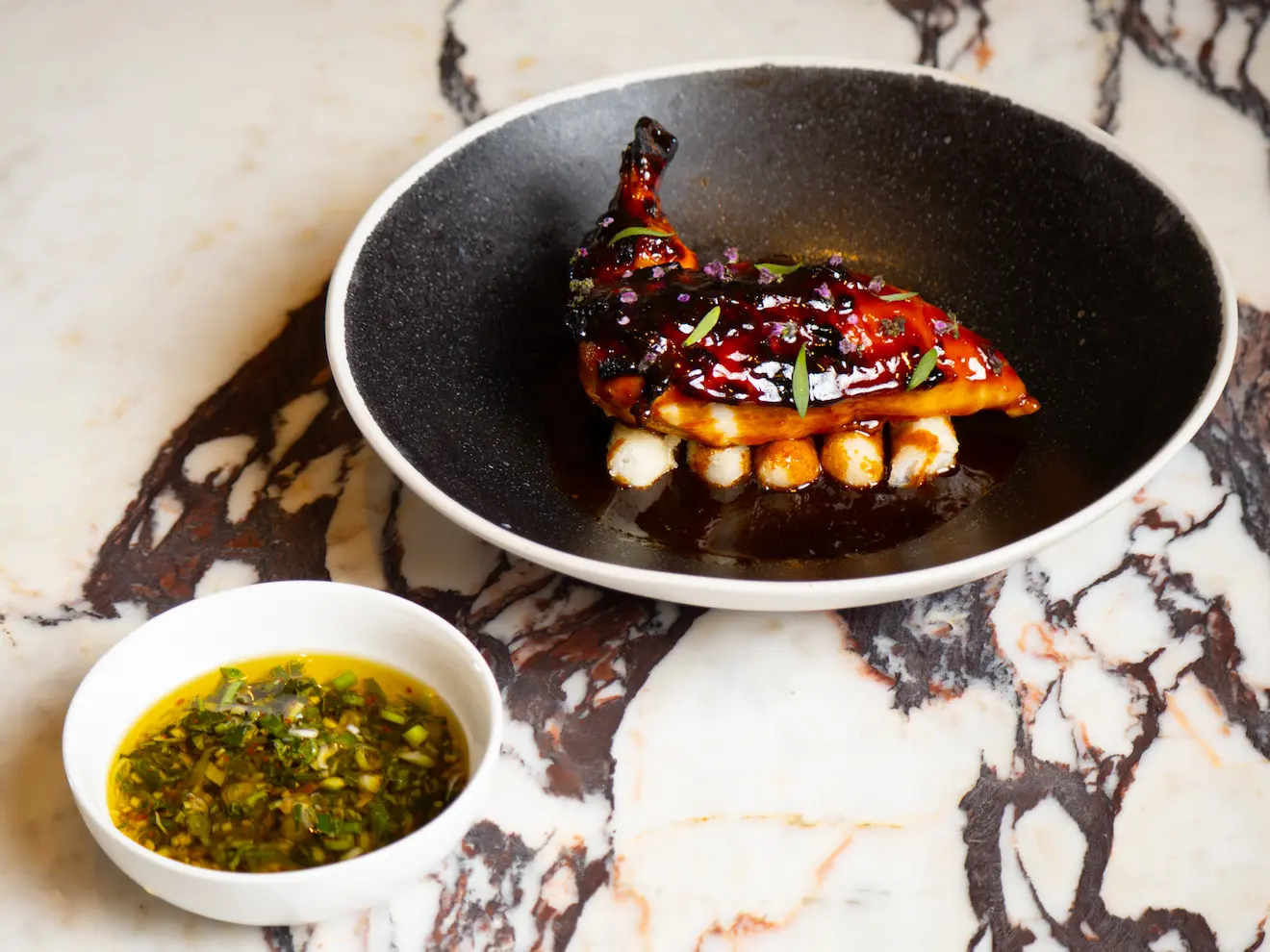 Gourmet roasted quail served over white beans with a side of herb sauce on a marble table - Spring Place