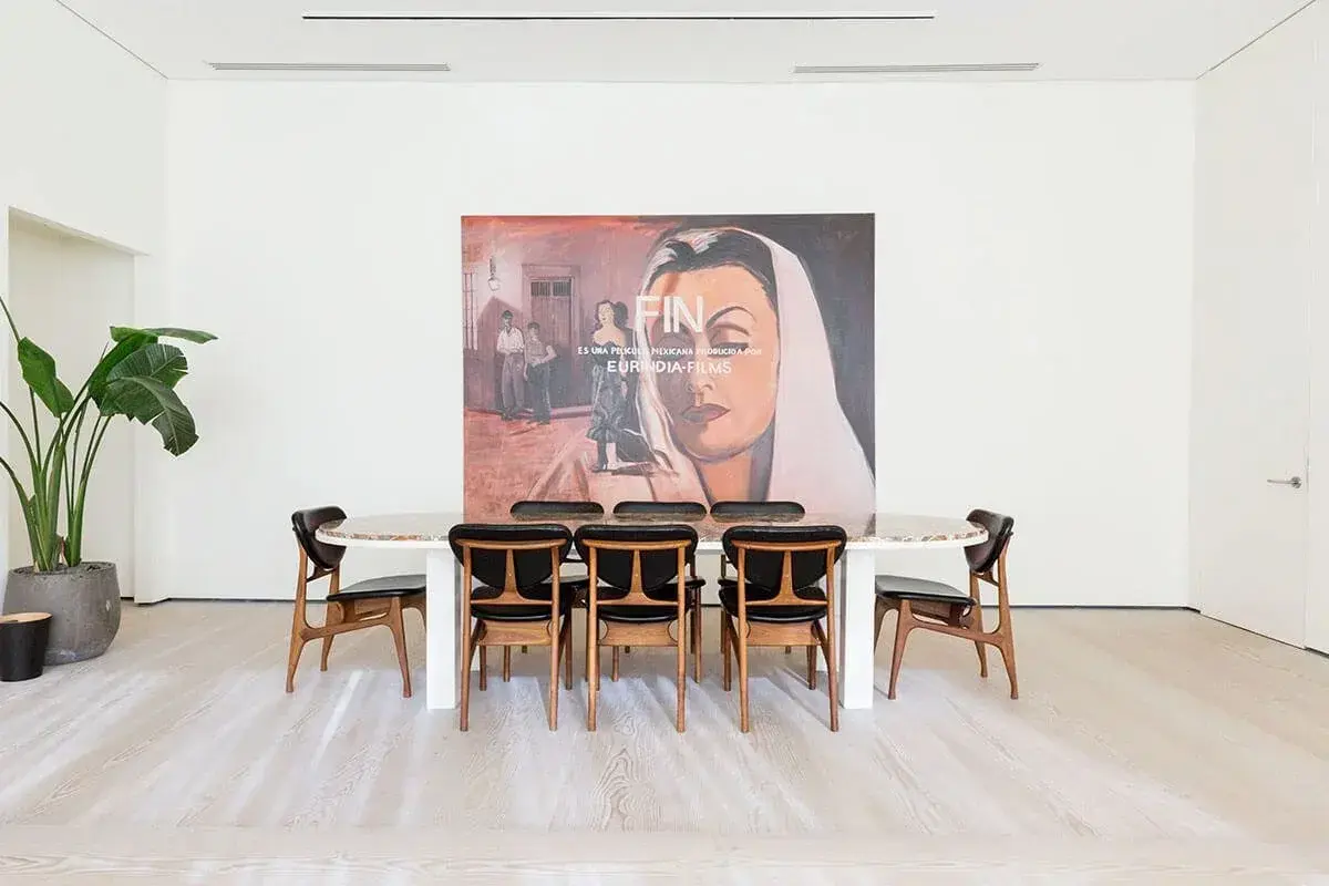 A modern room with a large painting of a woman, a long table, and chairs, with a plant in the corner.