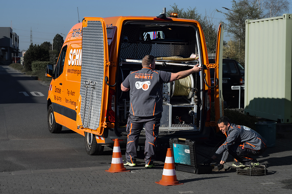 Zwei Mitarbeiter der Firma Rohrreinigung Schmitt arbeiten mit professionellem Equipment an einem geöffneten Einsatzfahrzeug. Einer bedient die Technik im Fahrzeug, der andere führt eine Kamera- oder Reinigungssonde in einen Kanalschacht ein. Im Vordergrund steht ein Absperrkegel zur Sicherung der Arbeitsstelle.