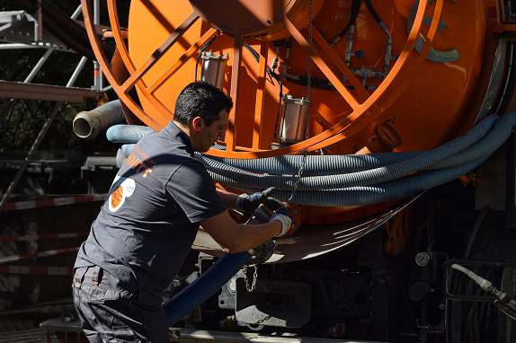 Ein Mitarbeiter der Firma Rohrreinigung Schmitt schließt einen blauen Saugschlauch an ein großes orangefarbenes Spezialfahrzeug an, das für die Entleerung von Gruben oder Abscheidern ausgestattet ist. Die Szene zeigt den professionellen Umgang mit moderner Technik im Einsatz.