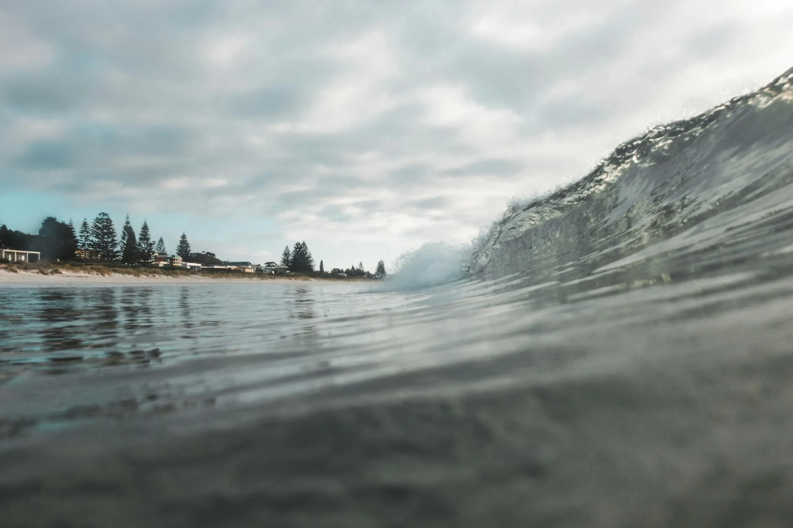 Close-up of a breaking ocean wave near a shoreline with trees and buildings under a cloudy sky.