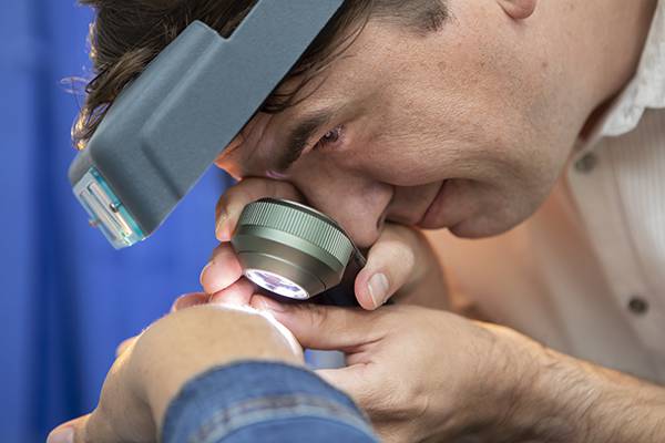 Dermatologist wearing a magnifying headlamp examining a patient's skin with a handheld dermatoscope.
