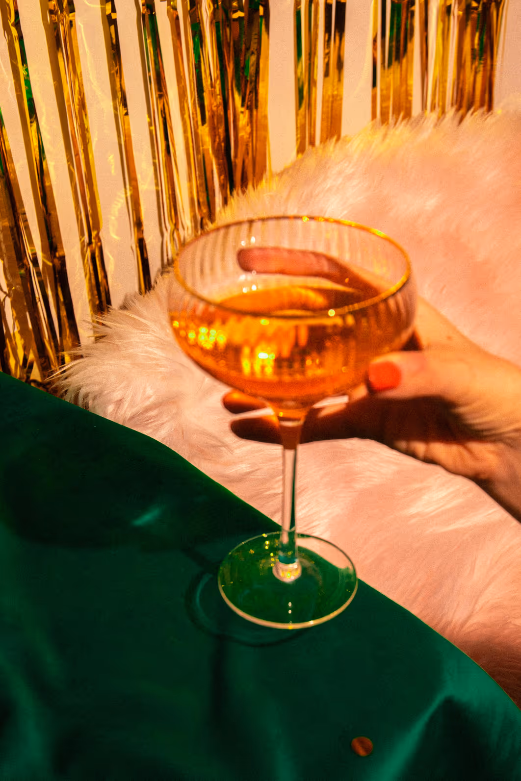 Hand holding an elegant coupe glass with a golden Madame Mocktail zero-proof drink at a celebration with gold tinsel backdrop