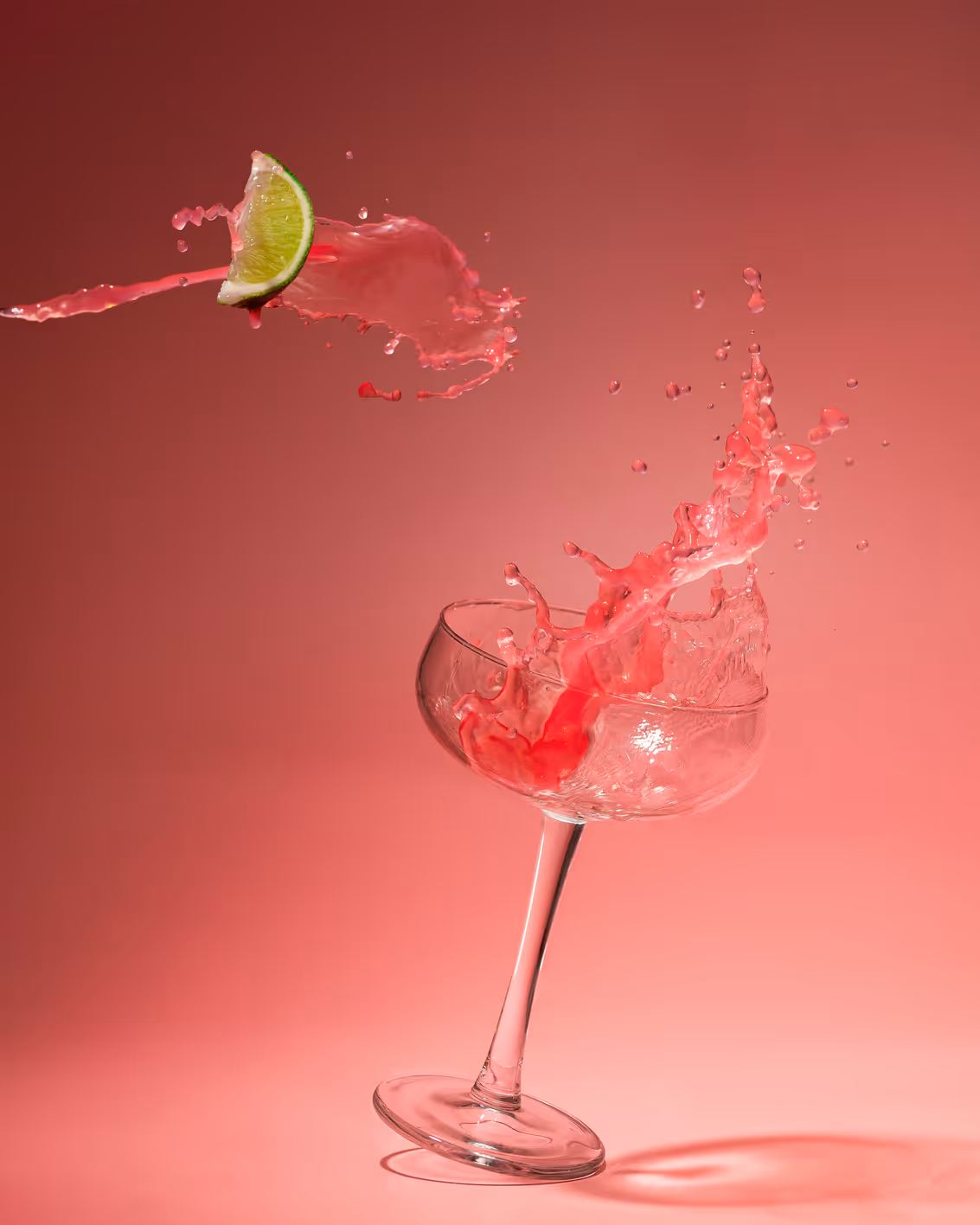 Vibrant cranberry Madame Mocktail zero-proof drink splashing dramatically from a coupe glass with fresh lime garnish on pink background