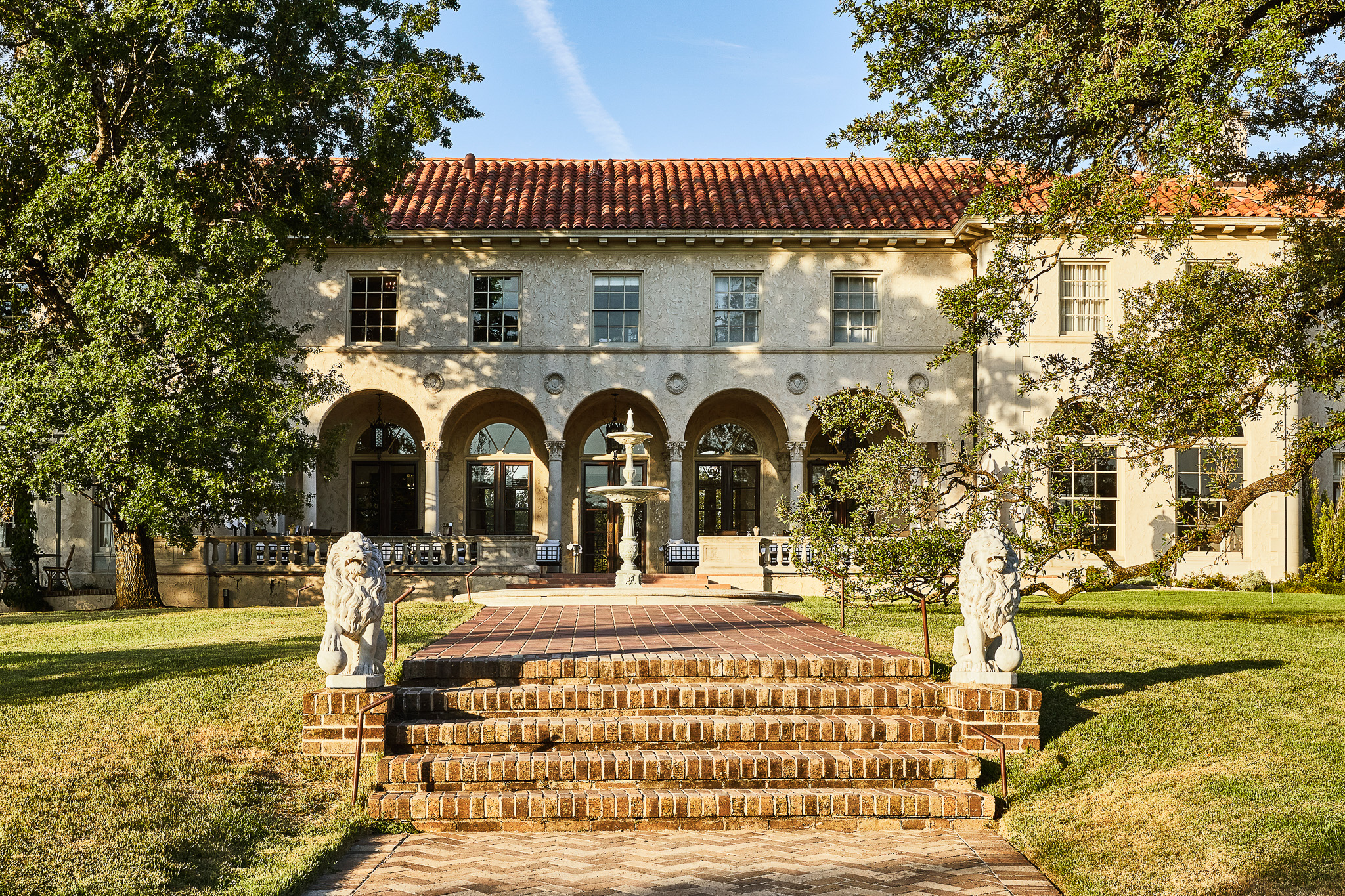 This image of the Commedore Perry Estate features Spanish roof tiling, textured stucco, and classic archways and adornments throughout with a brick pathway leading up to this magnificent structure.