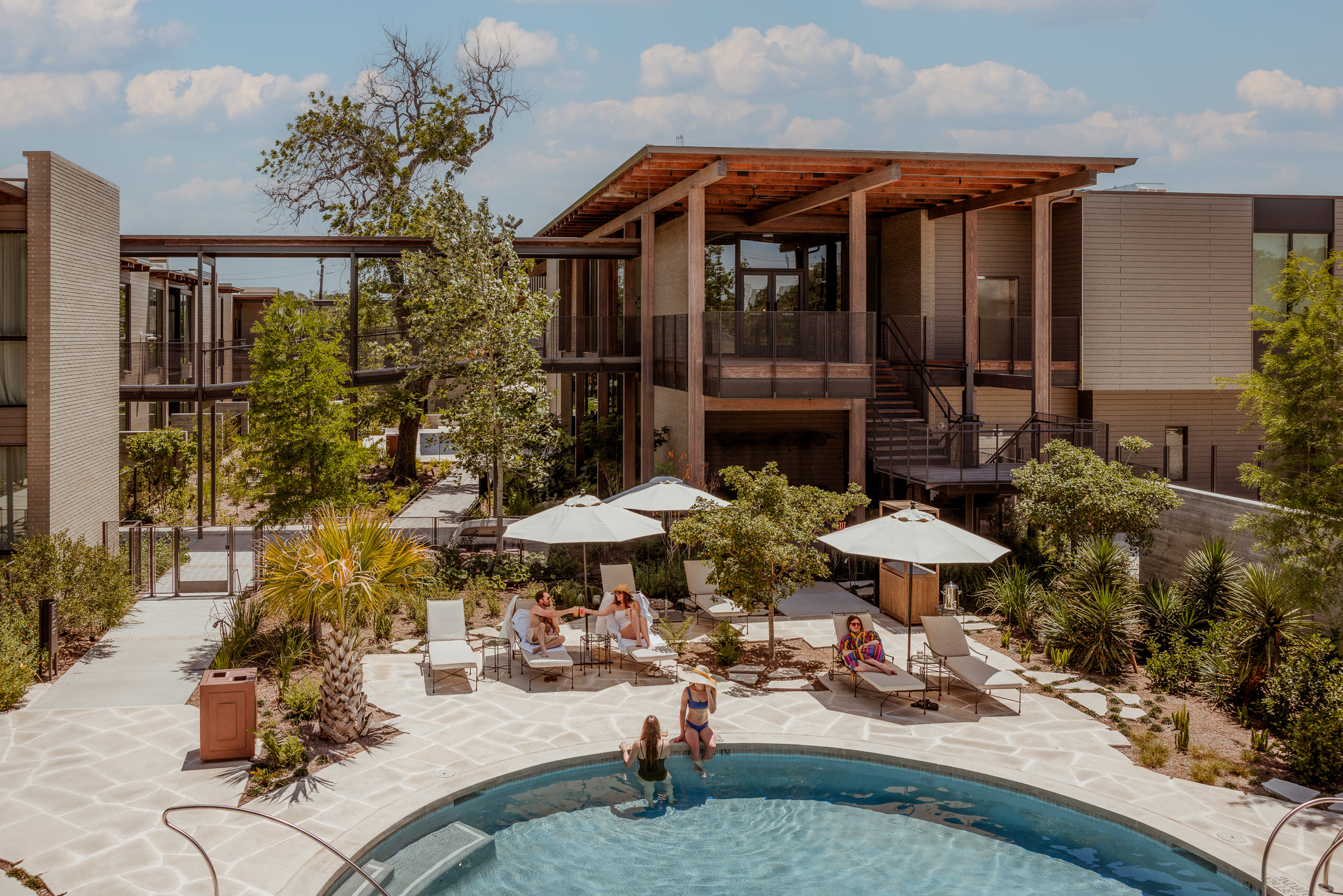 This image features the pool area of the Hotel Saint Augustine and a gorgeous array of plants and trees surrounding the space. Hotel guests lounge beneath the pool umbrellas and dip their feet in the pool.
