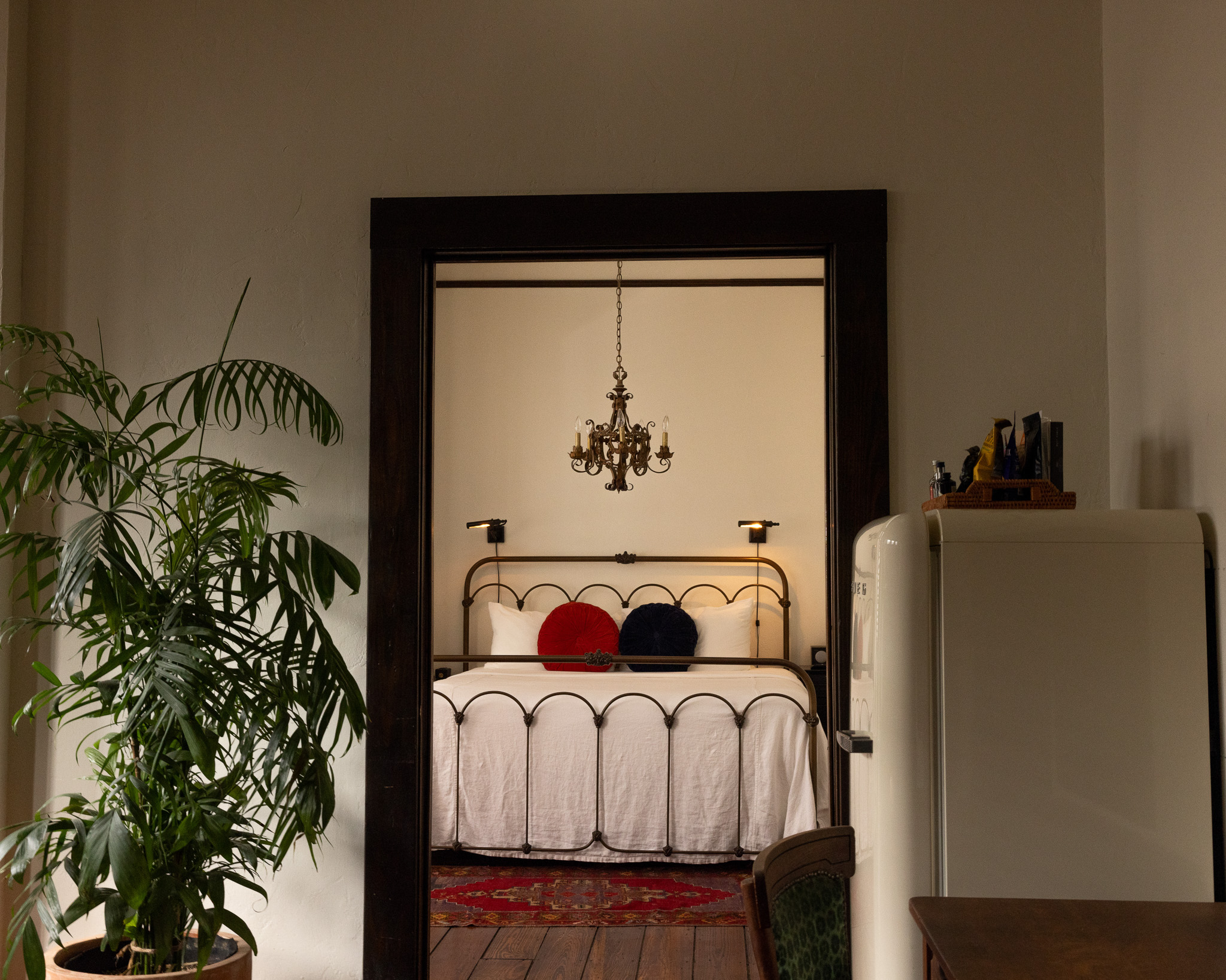 This iamge shows a shot in a room at the hotel Havana. There is a beautiful plant on the left, and retro cute fridge on the right, and the bedroom door frame centered in the shot, so one can see the bed therein.