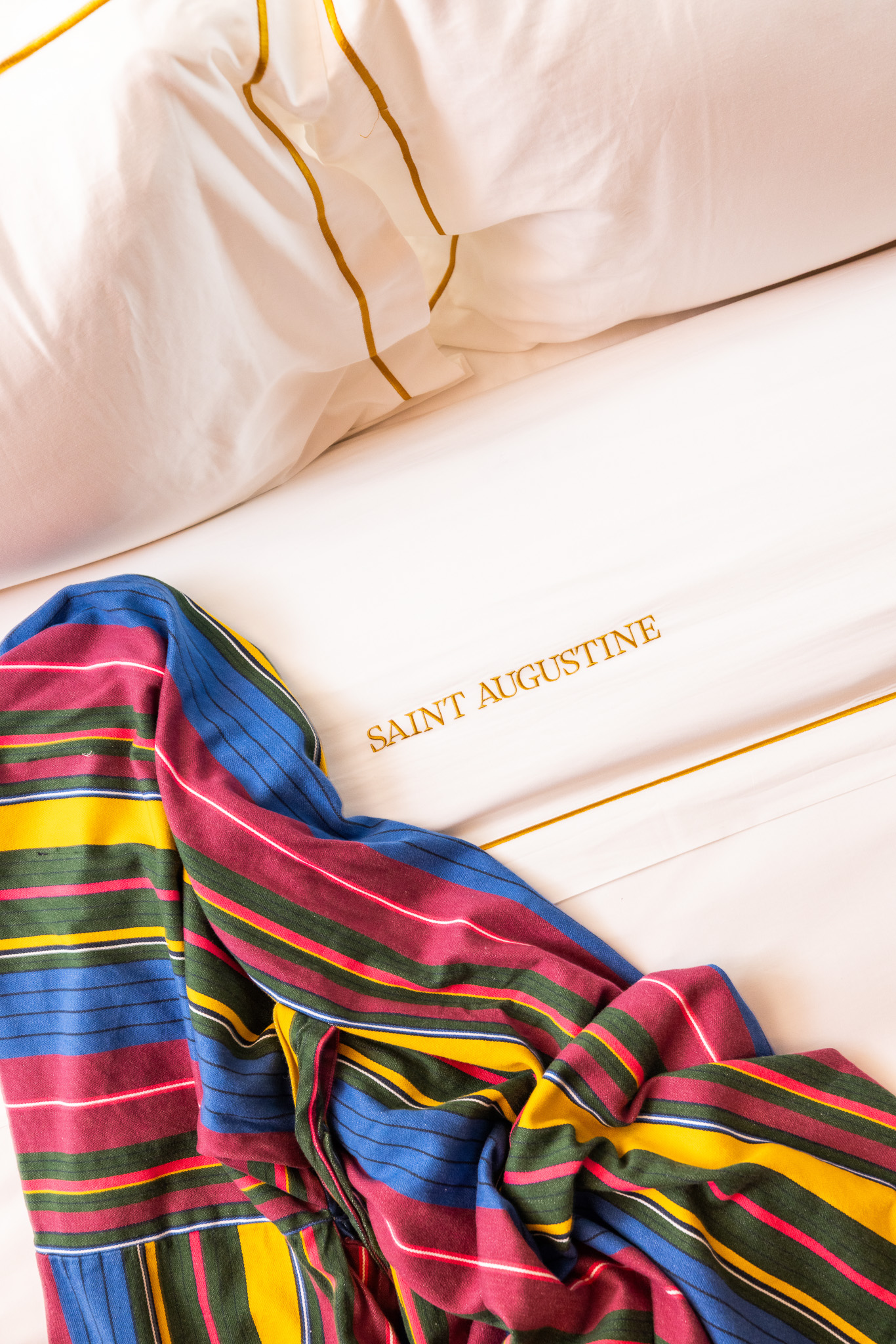 This image features the gold stitchign detail on the white sheets at the Hotel Saint Augustine with a colorful patterned throw blanket draped nearby.