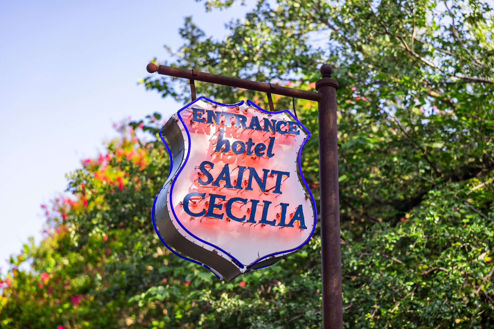 The sign to the entrance of hotel Saint Cecilia has neon red glow behind the navy blue lettering that protrude from this metal shield-shaped sign. It hangs from a vintage, rusty sign holder and has blue neon tubes that glow at night and outline the shield shape on both sides.