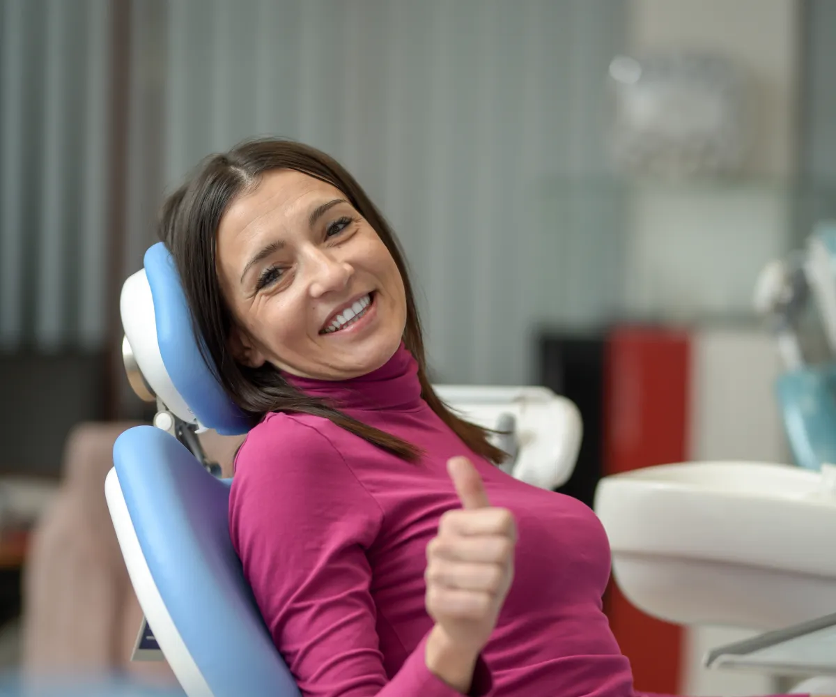 Woman sitting in a dental chair, smiling and giving a thumbs-up gesture.