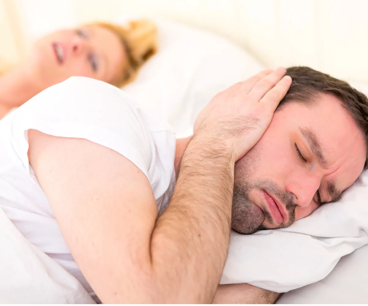 Man covers ears with pillow while trying to sleep next to a snoring woman.