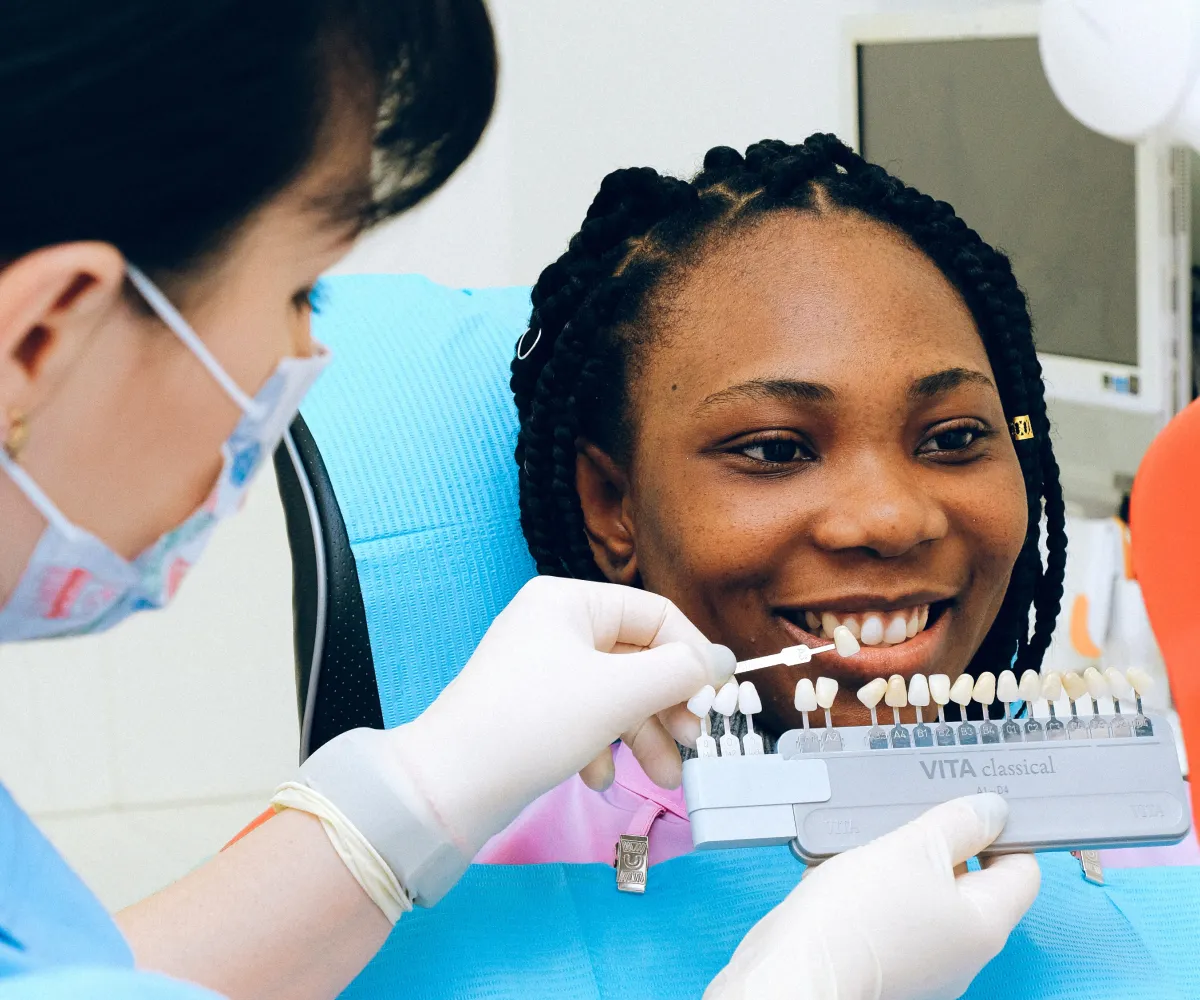A dental professional holds a tooth shade guide next to a smiling patient for color matching.