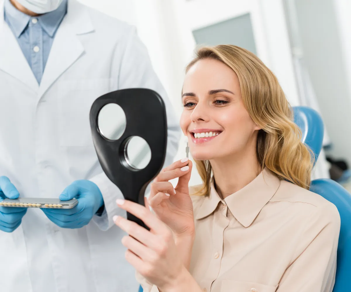A woman in a dental office holds a handheld mirror and inspects her teeth with a dentist nearby.