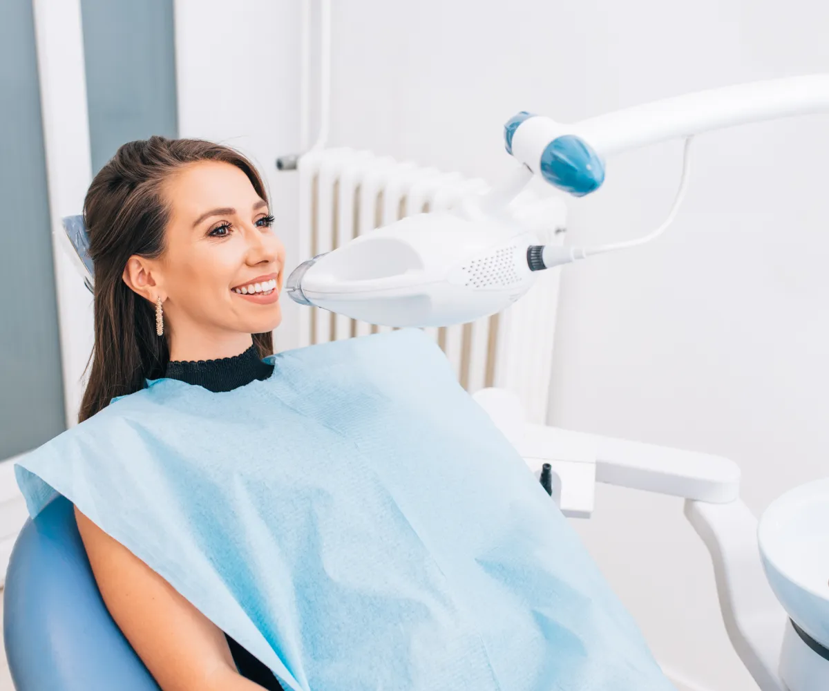 A woman sits in a dental chair receiving a teeth-whitening treatment under a blue light.