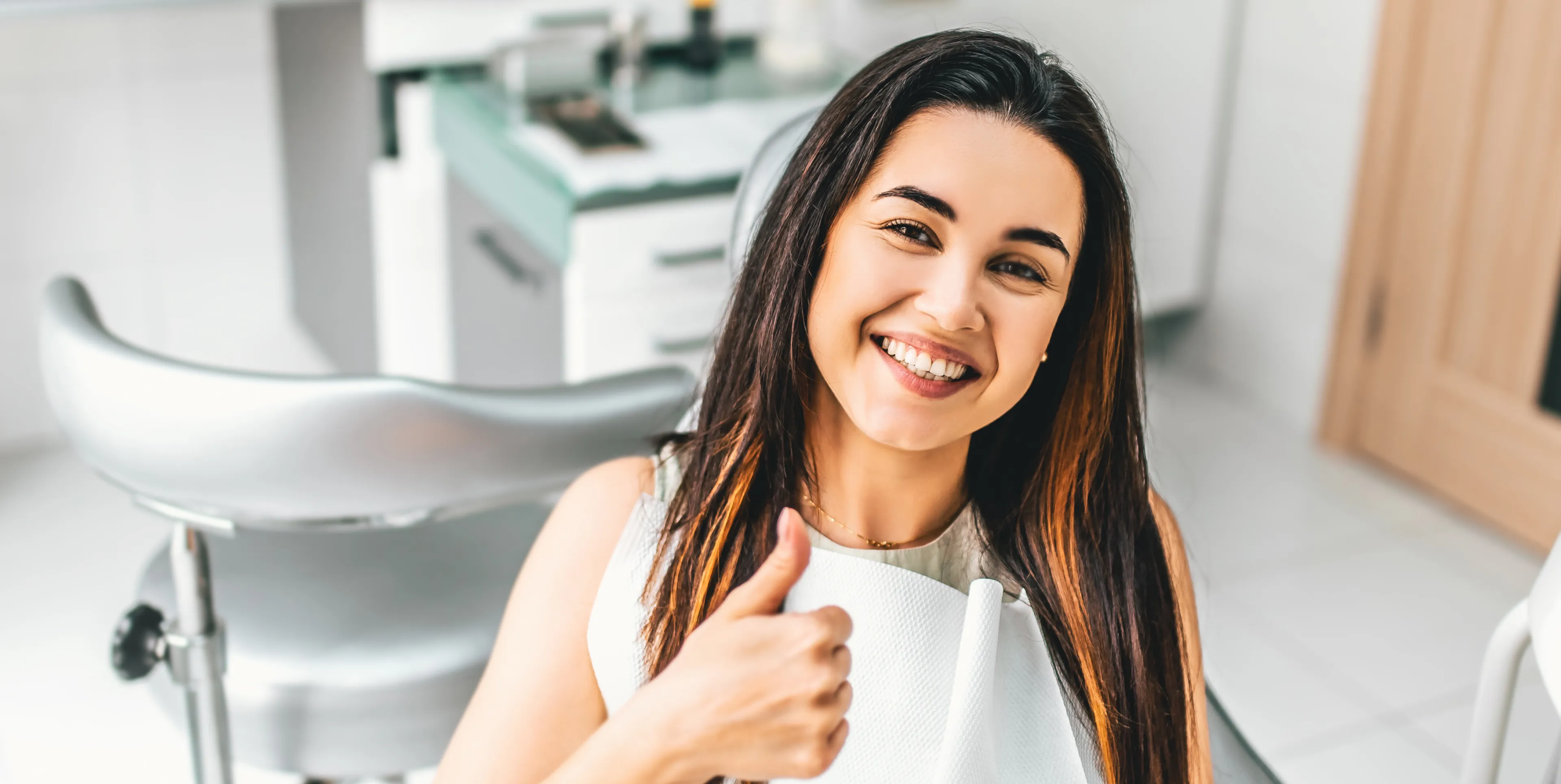 Smiling woman at the dentist gives a thumbs-up sign, sitting in a dental chair.